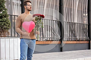 Young man with bouquet of roses and balloon waiting for his girlfriend outdoors