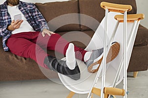 Young man with broken leg or injured foot sitting on couch and using mobile phone