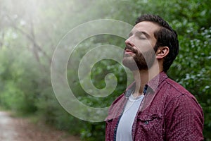 young man with brown hair and beard wearing red button down breathes in a breath of fresh air immersing himself in a nature path