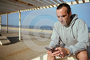 Young man browsing fitness app on mobile phone, scrolling newsfeed, checking social media content in Atlantic promenade