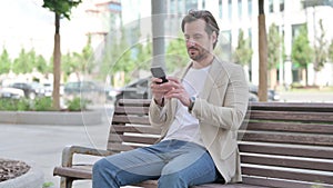 Young Man Browsing Internet on Smartphone while Sitting on Bench