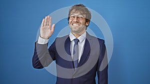 Young man in business suit joyfully saying hello, waving with a cheerful smile. friendly welcome gesture against isolated blue