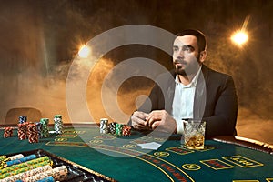 A young man in a business suit sitting at the poker table. Man gambles. The player at the gaming table playing cards.