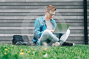 Young man businessman in shirt, jeans and glasses sitting on green grass and using laptop computer on the background of a gray