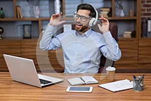 A young man businessman sits in the office at the table, works on a laptop in white headphones and listens to music