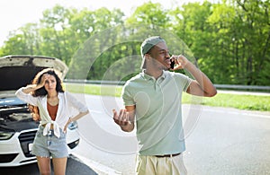 Young man calling for help with broken car, speaking to rodside assistance service, his upset girlfriend standing nearby