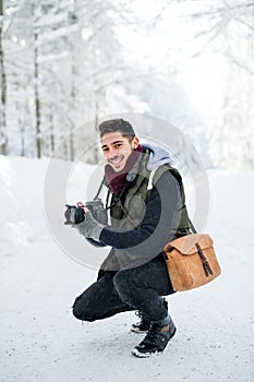 Young man with camera outdoors in snow in winter forest, looking at camera.