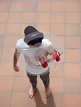 Young man with cap and headphones doing excercises weight training barefoot lifting arm with dumbbell on terrace outdoor