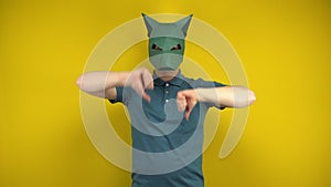 Young man in a cardboard jackal mask shows dislikes with thumbs on a yellow background. Man in a green polo and mask.