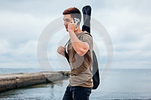 Young man carrying acoustic guitar and talking on phone walking on cloudy beach
