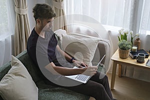 Young man in casual clothes sexing on comfortable sofa and surfing internet, shopping online on laptop computer