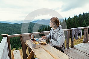 A young man in casual clothes sits at a table in a country house in the mountains, eating and looking away at the beautiful