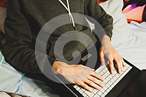 Young man in casual clothes sitting on an unmade bed working remotely from home with a laptop on his knees. Closeup shot