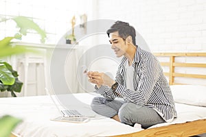 Young man in casual clothes and working at home while sitting on the bed,with a laptop in bedroom, play mobile games and chat on t