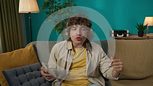 Young man in casual clothing and headphones sitting on the couch in the living room and listening to music on smartphone