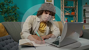 Young man in casual clothing, sitting on the couch in the living room and working on his laptop with focused face