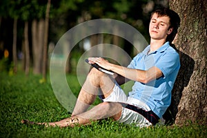 Young man sitting on green grass near tree and reading book