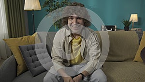 Young man in casual clothing, sitting on the sofa in the living room looking at the camera and smiling HDR BT2020 HLG