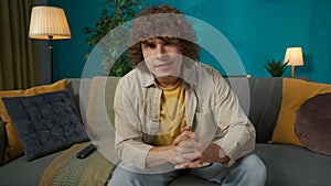 Young man in casual clothing, sitting on the sofa in the living room and watching news report on tv