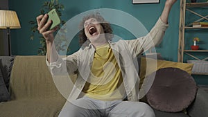 Young man in casual clothing, sitting on the sofa in the room with a bootball ball and holding a smartphone in hand HDR