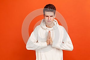 Young man in casual style hoodie pressing hands together showing namaste gesture, practicing yoga with peacefull face and closed photo