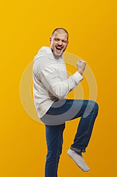 Young man celebrating success, wearing white hoodie and jeans raising fist up and leg