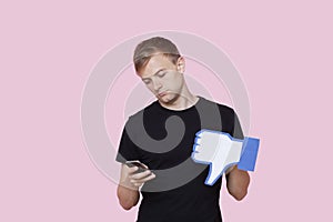 Young man with cell phone holding fake dislike button against pink background