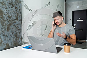 Young man with cell phone working on laptop at table in the kitchen