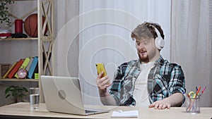 Young man in checkered shirt typing on laptop keyboard in big white headphones, then talking on video call on phone