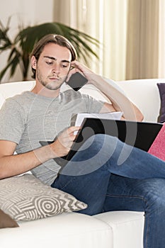 Young man checking some notes, holding his phone and a laptop while sitting on a sofa