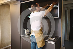 Young man in a chef's apron in a home kitchen taking out plates from the kitchen cabinet