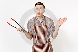 Young man chef in striped brown apron, shirt holding black kitchen serving plastic pair of tongs for salad isolated on