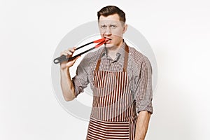 Young man chef in striped brown apron, shirt holding black kitchen serving plastic pair of tongs for salad isolated on