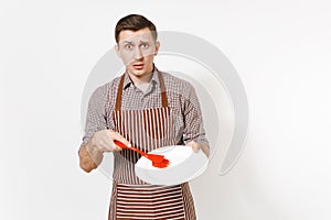 Young man chef in striped brown apron, shirt holds washes white round empty clear plate with red brush for washing