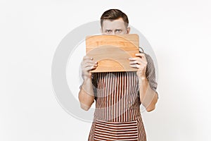 Young man chef or waiter in striped brown apron, shirt cover face with wooden cutting board, knife isolated on white