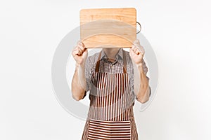 Young man chef or waiter in striped brown apron, shirt cover face with wooden cutting board, knife isolated on white