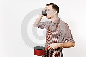 Young man chef or waiter in striped brown apron, shirt holding and tasting red empty stewpan black ladle isolated on