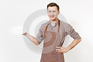 Young man chef or waiter in striped brown apron, shirt holding white round empty clear plate isolated on white