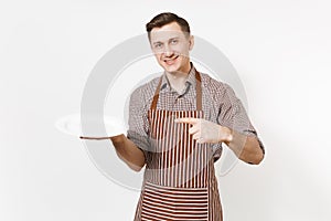 Young man chef or waiter in striped brown apron, shirt holding white round empty clear plate isolated on white