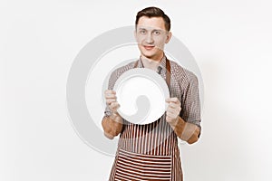 Young man chef or waiter in striped brown apron, shirt holding white round empty clear plate isolated on white