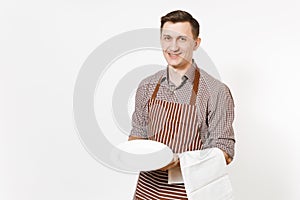 Young man chef or waiter in striped brown apron, shirt holding white round empty clear plate, towel napkin isolated on