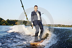Young man in classic suit rides wakeboard on a lake near city holding halyard. Happy clerk escaped from stuffy office to