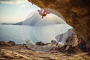 Young man climbing challenging route in cave at sunset. His partner belaying him