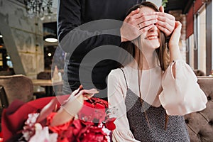 A young man closes his eyes to the girl, his wife, and gives a bouquet of red flowers in a cafe by the window.