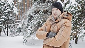 Young man in coat posing with hands crossed and smiling at camera on background of snowy forest.