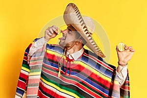 Young man in colorful poncho and sombrero squeezing lime on his tongue, posing against yellow studio background