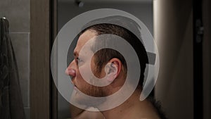 A young man is combing his hair in the bathroom while standing in front of the mirror. A man with a beard with a comb in his hands