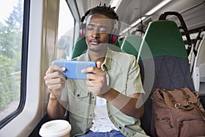 Young Man Commuting To Work Sitting On Train Streaming And Watching Film Or Show On Mobile Phone