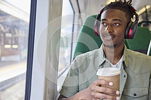 Young Man Commuting To Work Sitting On Train Wearing Wireless Headphones With Takeaway Coffee