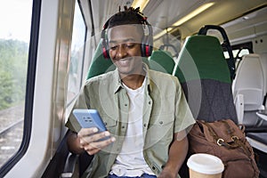 Young Man Commuting To Work On Train Sitting On Train Looking At Mobile Phone For Travel Information Or Social Media